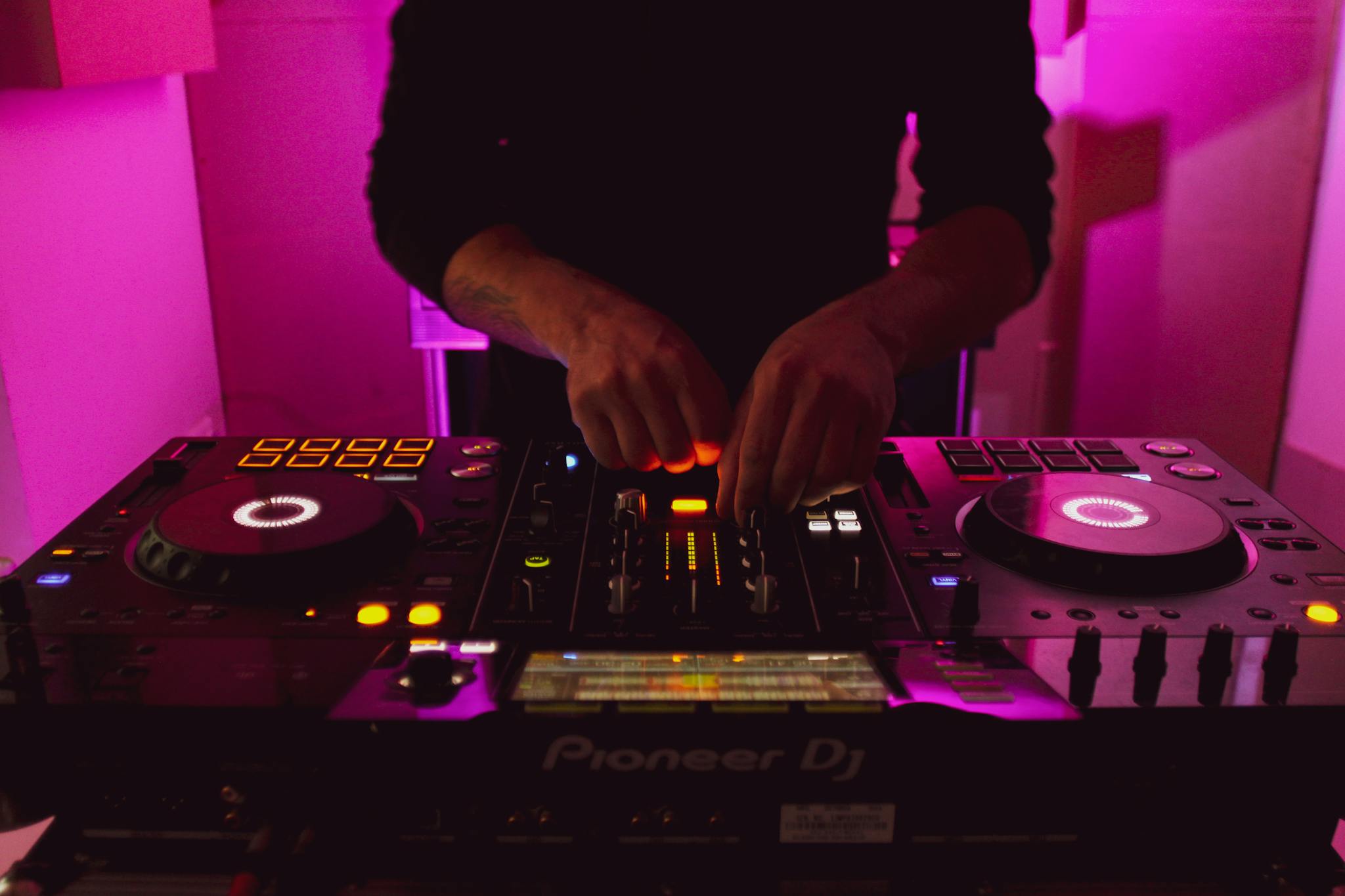 Close-up of DJ hands mixing music on a Pioneer DJ controller with vibrant pink lighting.